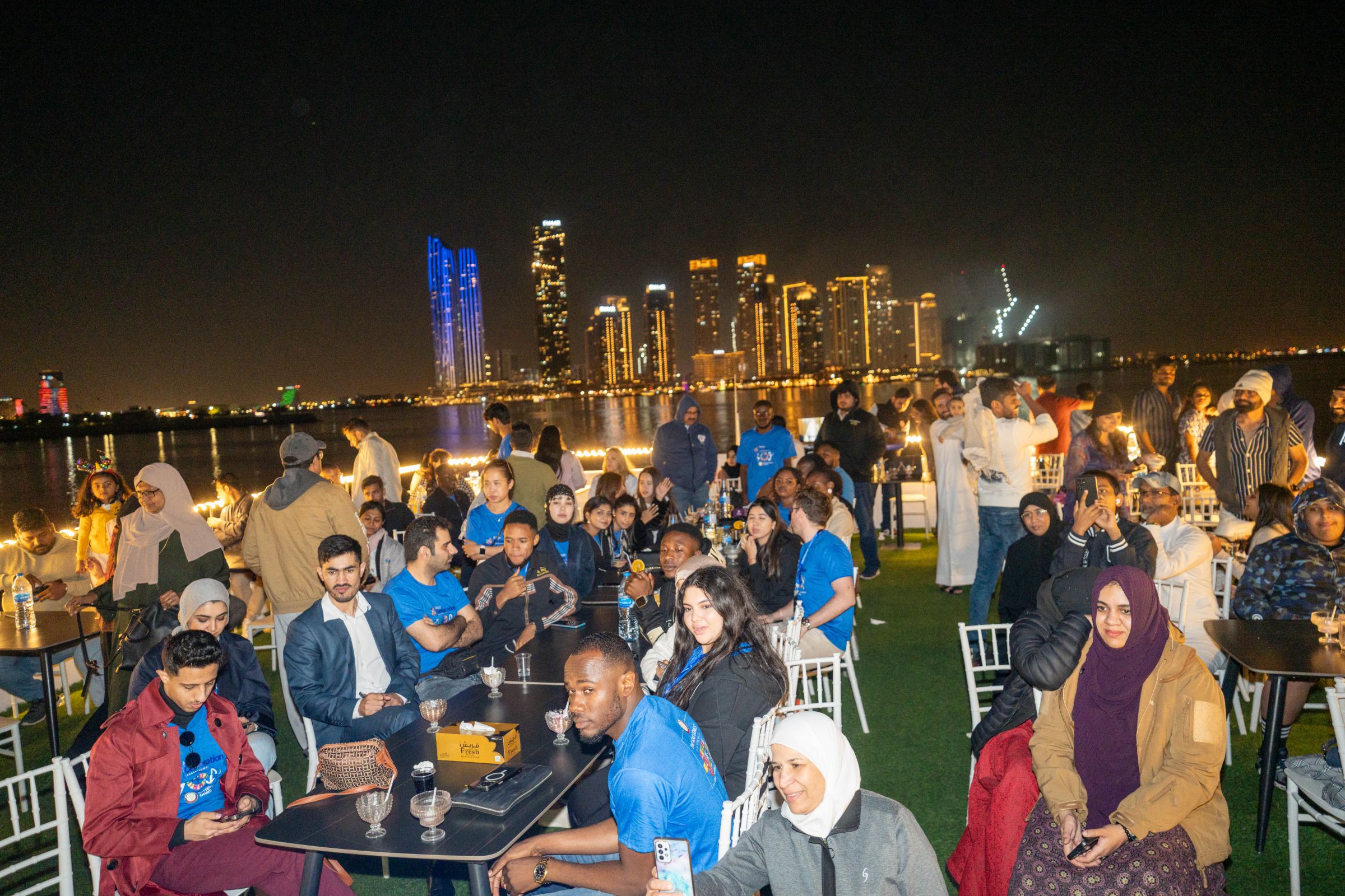 Delegates seated at tables on a lawn by the water at night with the Dubai skyline behind them