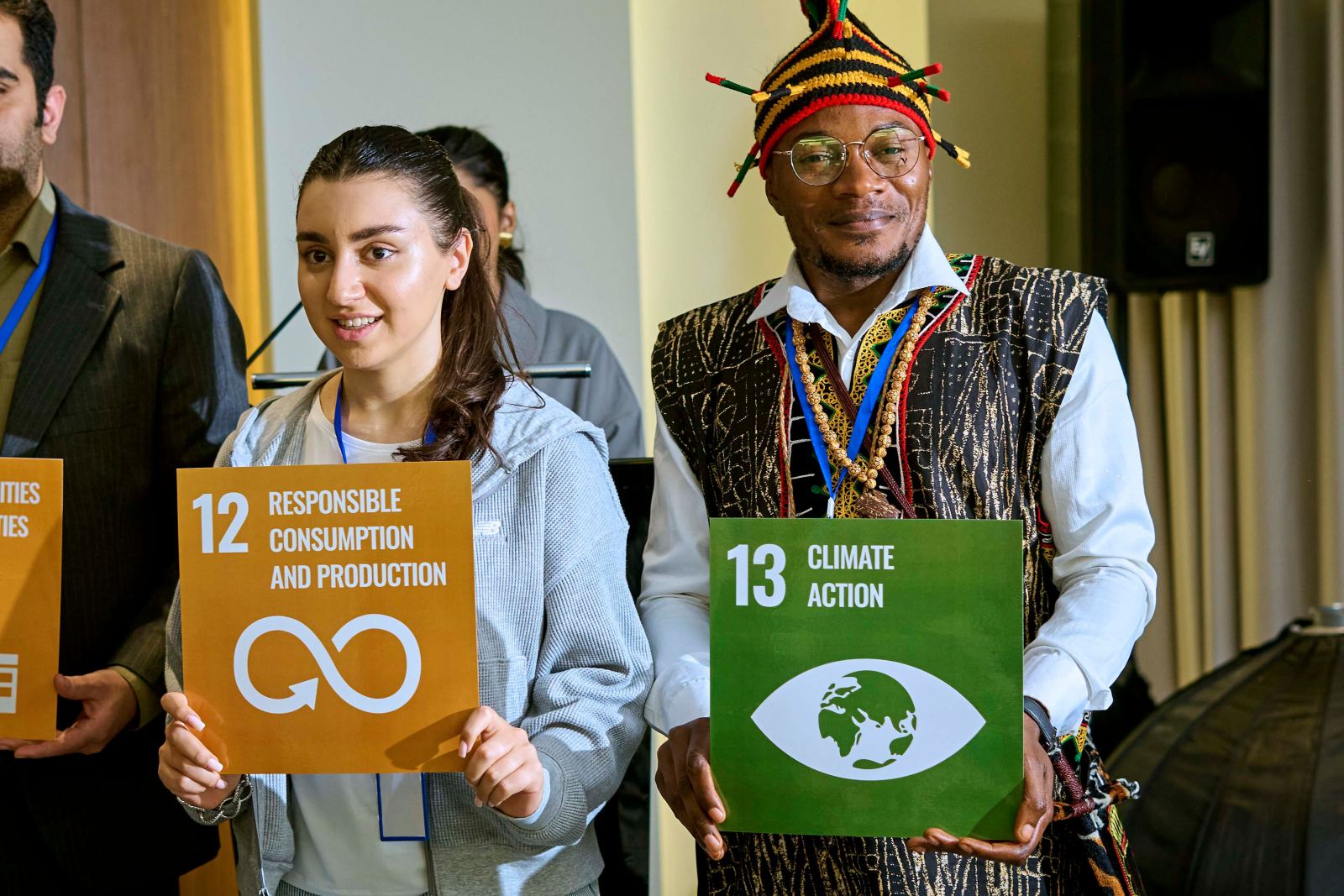 Two delegates holding Sustainable Development Goal cards 12 and 13, one wearing traditional dress