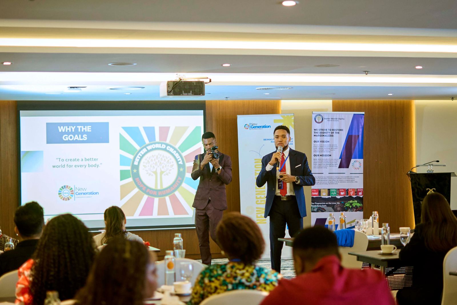 A speaker presenting to delegates in front of a slide titled Why the Goals